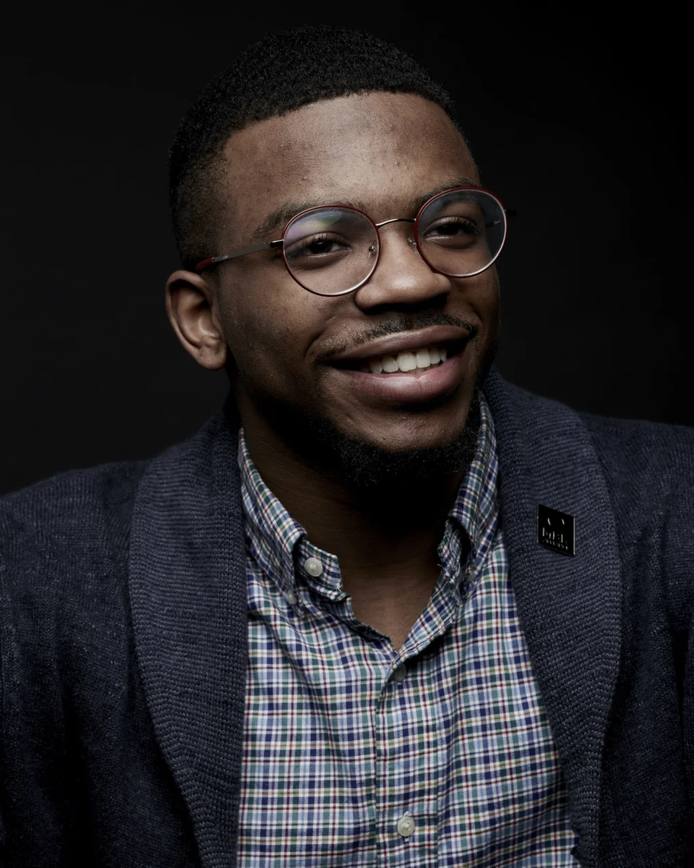 A Black man with short hair and glasses smiles slightly as he looks to the right. He wears a plaid button-down shirt and dark jacket. The background is black and the lighting is soft on his face.