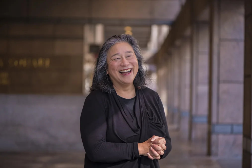 A photo of a woman, mid-laugh with her hands clasped in front of her. She has gray and black hair and wears a draped top. She stands in a large room with columns.  
