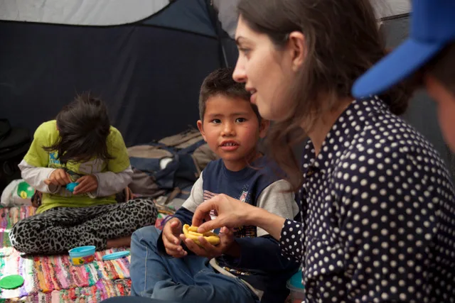 The camera focuses on a young boy with a light-medium skin tone sitting in a tent while holding snacks. Surrounding him is a young girl with a light-medium skin tone and a woman with a light skin tone