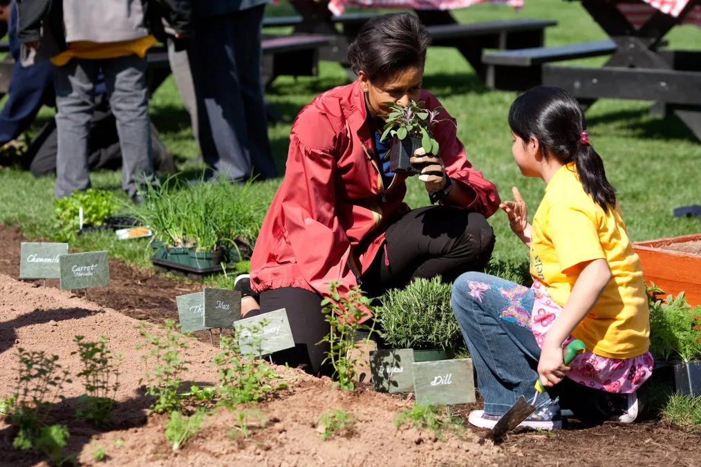 
First lady, Michelle Obama kneels down while sniffing a plant in a garden while a young girl with a light skin tone kneels in front of her 