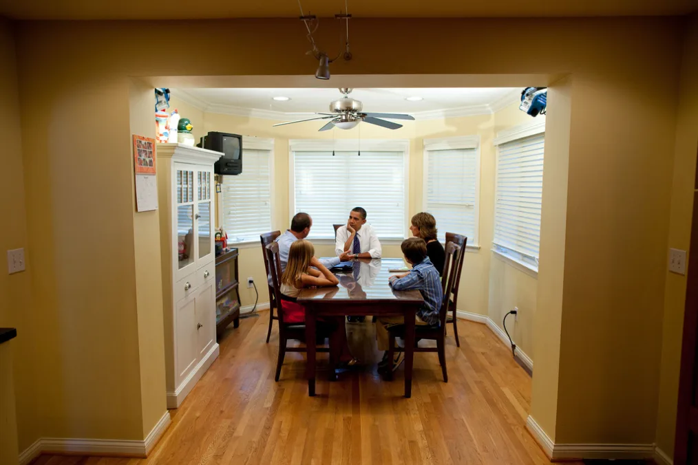 President Obama sits at a kitchen table with a family with a light skin tone.