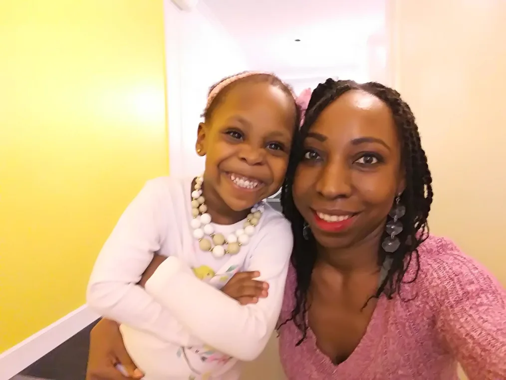 A picture of a woman with shoulder length twist wearing a pink shirt and her daughter who is wearing a white long sleeve smiling and they're both medium brown skin