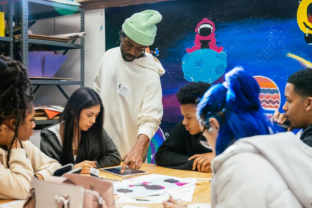 Brandon Breaux, a Black man with a deep skin tone, stands over five seated students. On the table is an iPad and illustrations. He is wearing a mint beanie. Space art is on the wall in the background.