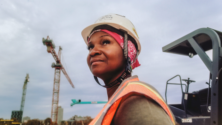 A Black woman wearing a hard hat and orange safety vest looks off into the distance. There are cranes on the horizon behind her.