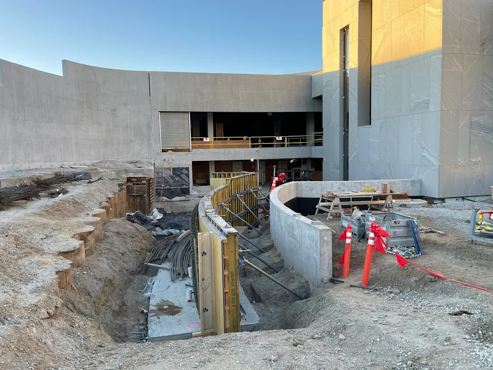 A construction site with concrete walls surrounding a large dug out area in the ground. The beginnings of walls are being propped up in the ditch with angled poles.