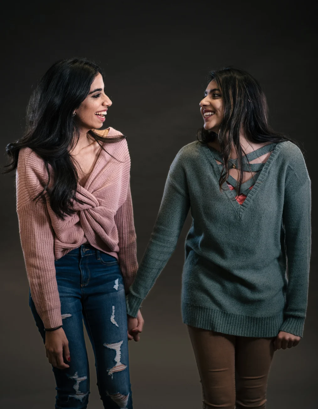 Two women with light-medium skin tones and long black hair hold hands while
smiling at each other in front of a black backsplash.