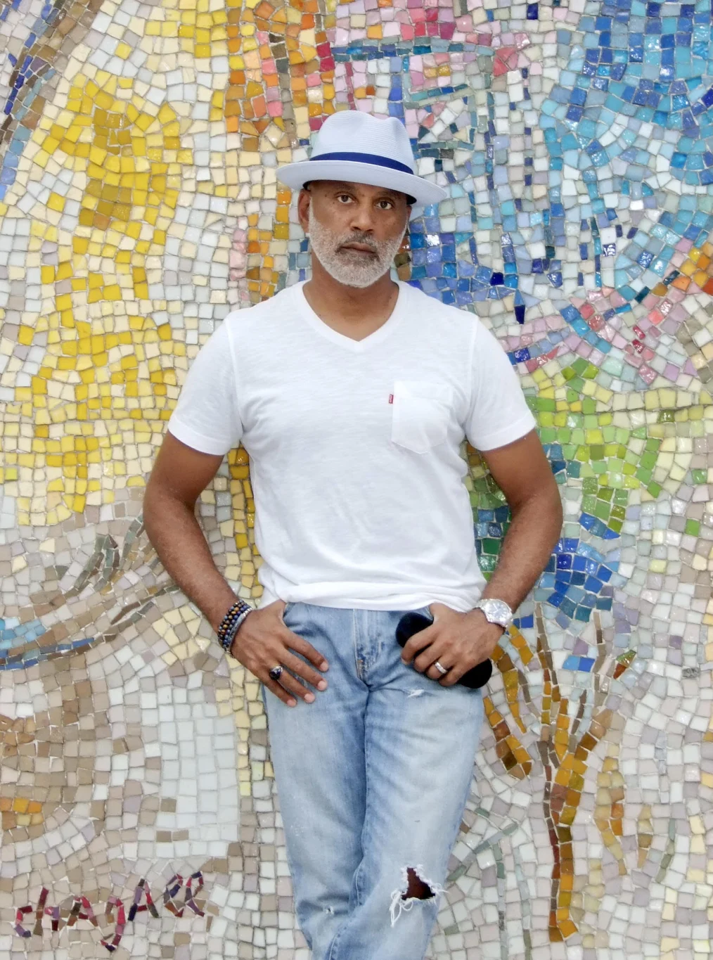 A man with a medium skin tone in a white fedora hat and a white tshirt stands in front of a colorful mosaic wall looking toward the camera.