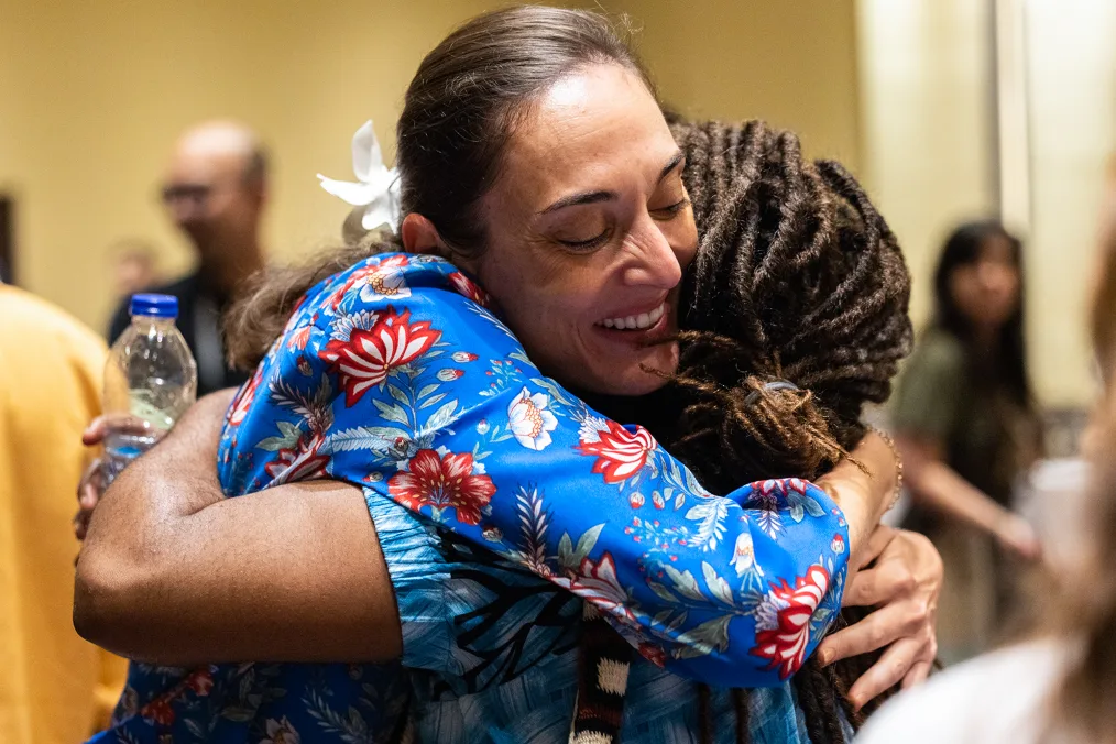 Two women embrace. One woman, facing the camera, has a medium skin tone and is smiling as she embraces a woman with a deep skin tone and brown locs. People are blurred in the background. 