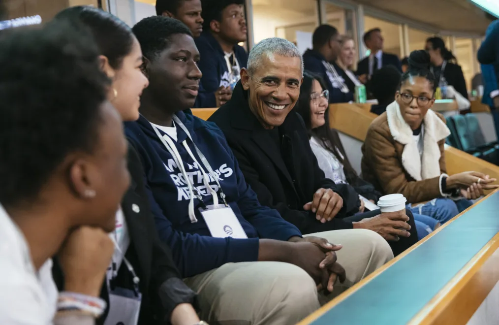President Obama is shown sitting beside a deep-skin-toned young man while smiling a talking to
a medium-skin-toned young lady. He is also surrounded by many other various skin-toned people.