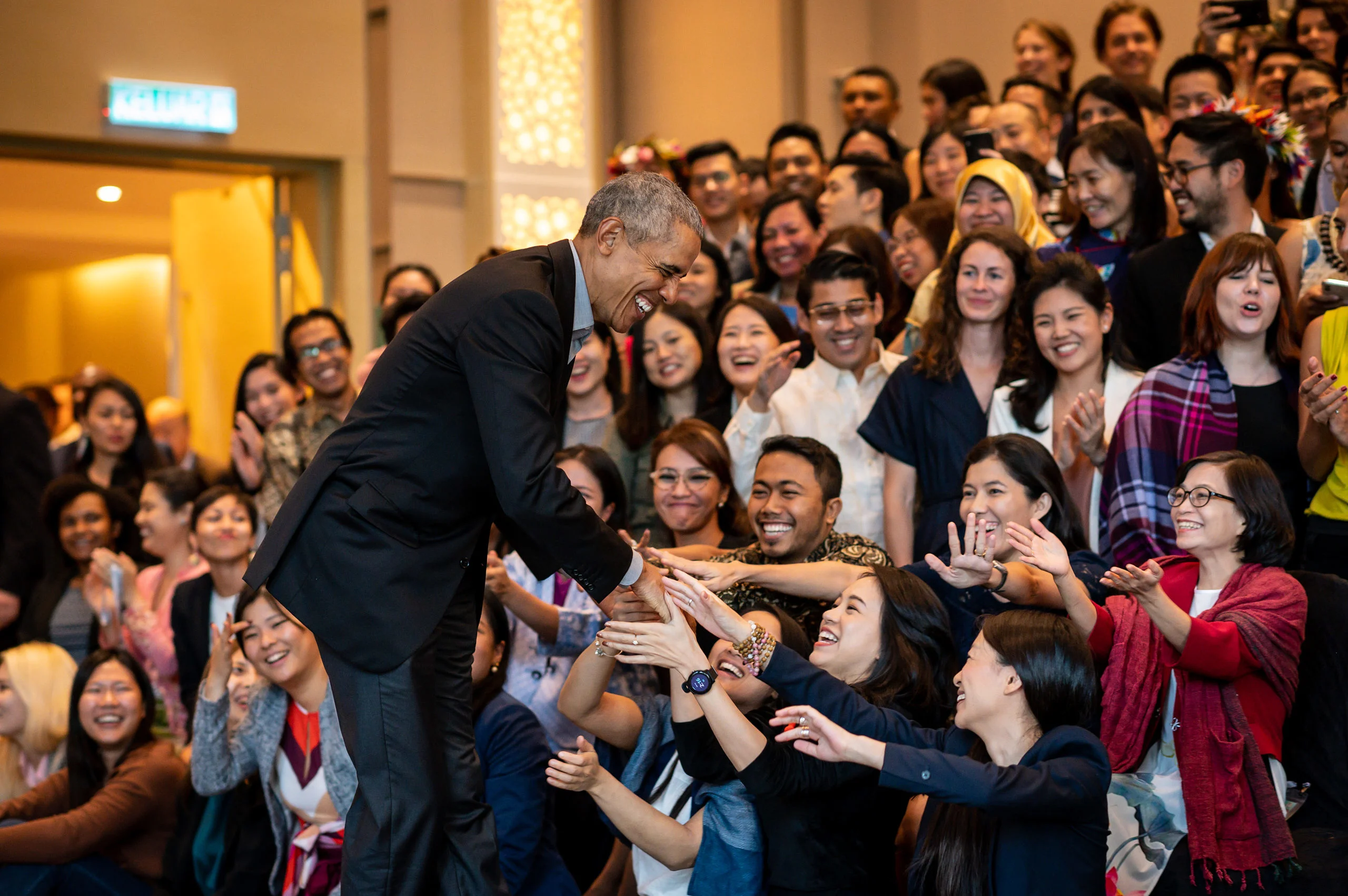 President Obama smiles as he shakes hands with an excited crowd of young people extend their hands. They have a range of light to medium skin tones. 