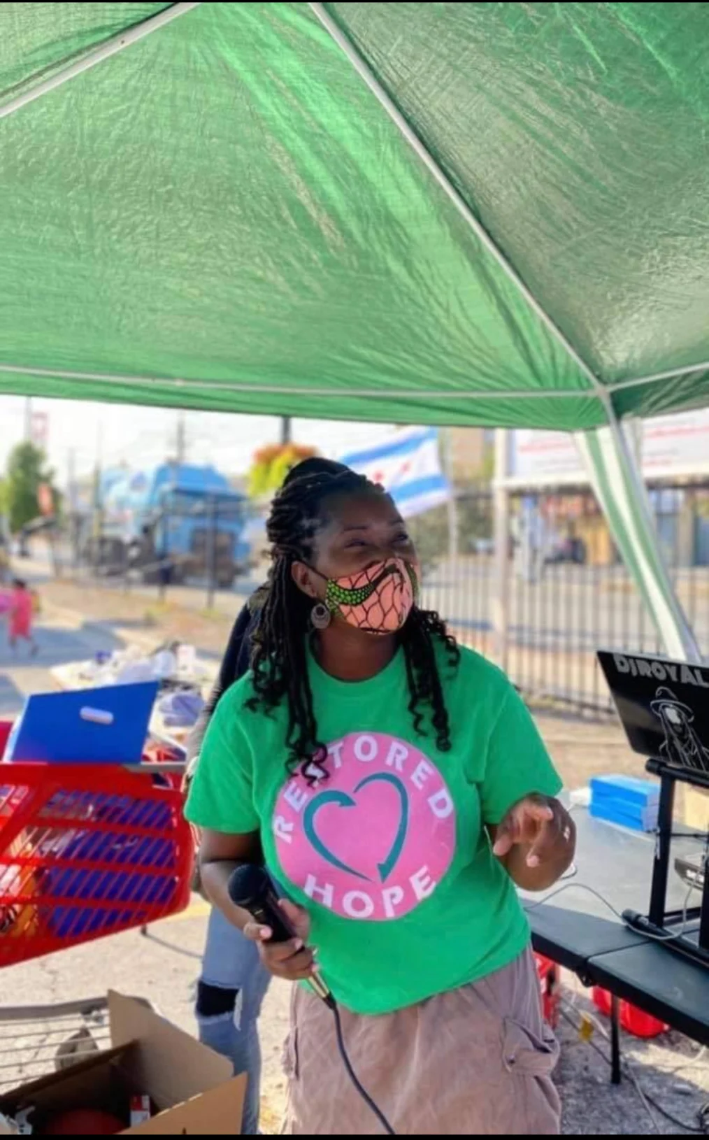 A Black woman with a deep skin tone stands  underneath a large green umbrella on a boardwalk. She has dreadlocks that hit past her shoulders and is wearing a green and pink shirt that reads, “Restored Hope.” She wears a patterned face mask and holds a microphone. Behind the woman is a red shopping cart, a stack of blue signs, and a DJ set up.