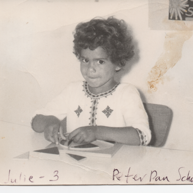 In a black and white photo, a young Julie Mehretu, plays with triangle blocks at a table. She is looking up at the camera. 