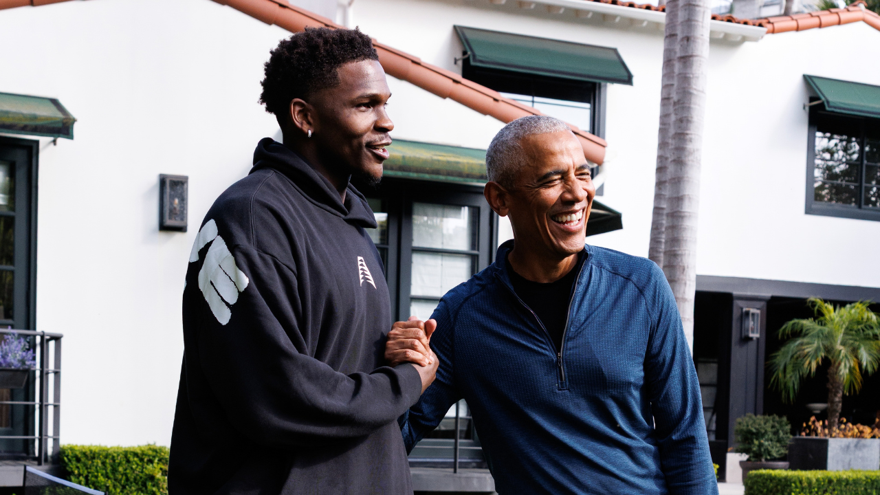Anthony Edwards and Barack Obama give each other a hand shake, more specifically a dap, as they stand next to each other. Anthony has a deep skin complexion short coiled hair and a goatee. He is wearing a black sweatshirt. Barack Obama has medium deep skin tone and closely cropped gray hair. He is wearing a light colored button down shirt.