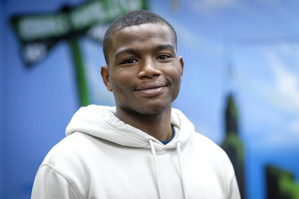 A Black man smiles at the camera. He wears a white hoodie and stands in front of a wall showing green and blue graphics.