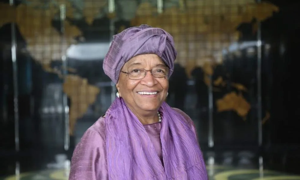Ellen Johnson, a black woman with a light-medium skin tone and thin metal glasses wearing a purple headwrap and matching scarf and dress, smiles towards the camera
