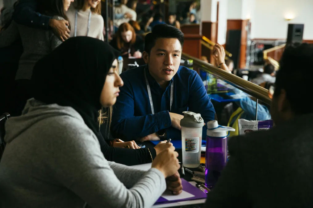 A man with a light skin tone wearing a blue button-up shirt sits at a cluttered table with a woman with a light skin tone wearing a gray hoodie and black hijab. There are other people blurred out at the table and surrounding the table. 