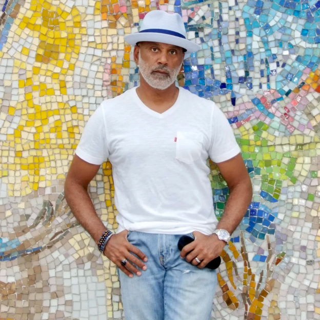 A man with a medium skin tone in a white fedora hat and a white tshirt stands in front of a colorful mosaic wall looking toward the camera.