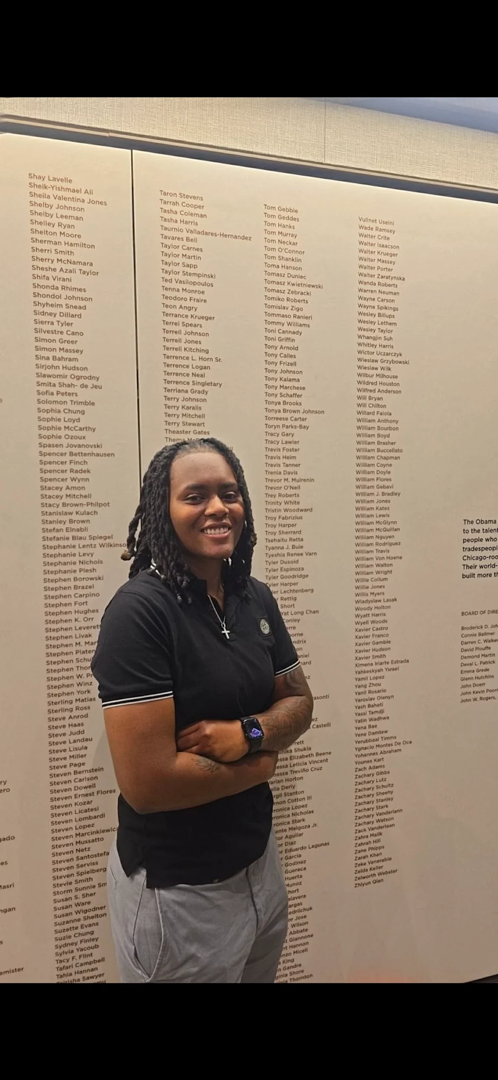 Toryn Parks, a Black woman with a medium skin tone and locs, stands next to her name on the worker wall at the Obama Presidential Center. She is wearing a navy polo and gray slacks.