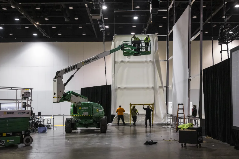 An image of the construction site of the Power of Words display at the Obama Presidential Center Museum. It is a wide shot of the Power of Words display and features a four story silver metal frame. Two construction workers with light skin tones are on a green crane hanging white canvas on the silver metal frame. On the floor are three people with light skin tones stand at the bottom of the crane under a scaffolding.
