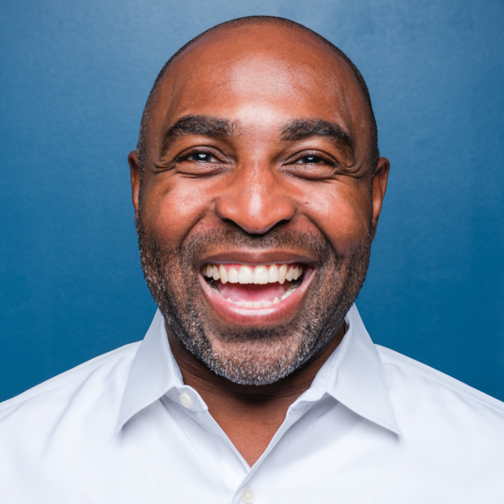 The image is a headshot of Clarence Fluker. He has a deep medium skin complexion, a bald head, brown eyes, a mustache, and a closely cut goatee. He is wearing a white button down shirt and smiling into the camera. The background behind him is blue.