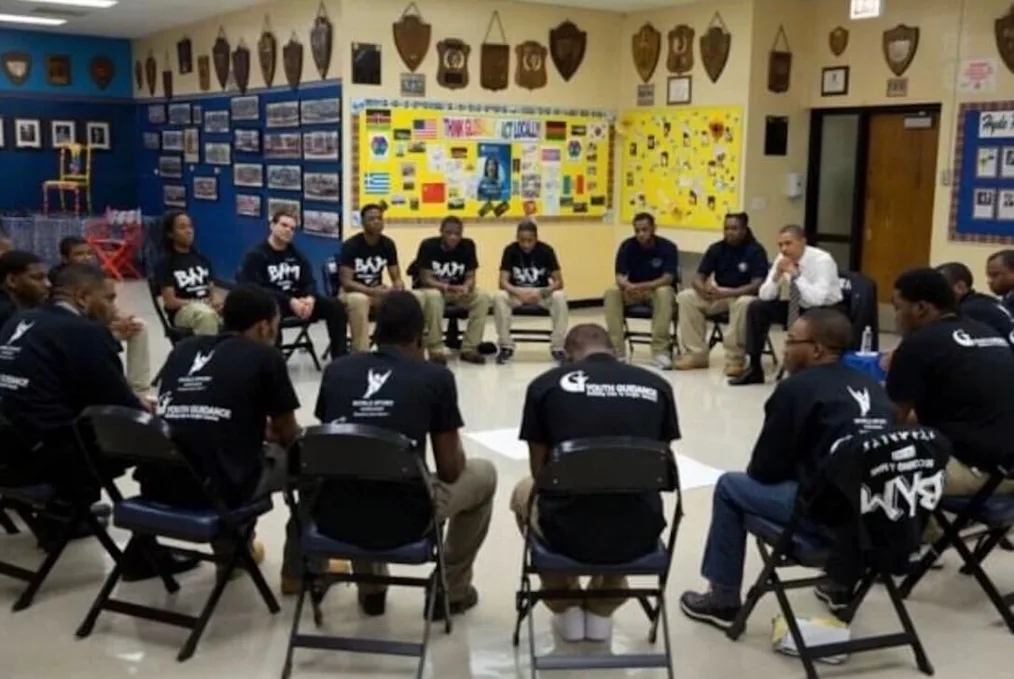 A group of young men with a range of light to dark skin tones sit in a circle with President Obama. President Obama is wearing a shirt and tie, Some wear shirts have white text that reads "BAM.” The walls behind them are decorated with a bulletin board that includes flags and other posters.