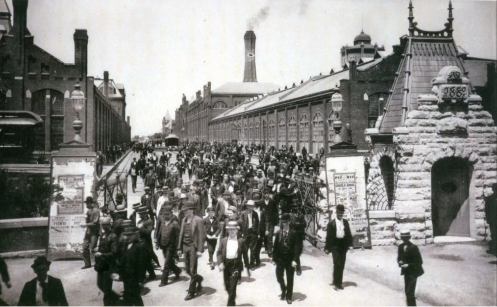 A old black and white photo depicting a crowd of men and woman in business attire walking through a gate. The metal gate supported by two brick structures on both sides. On top of the structures are streetlamps. In the right side of the picture is a gate management building. At the top of the building reads "1889". In the back ground are trolleys, a road, more people, and long industrial buildings.