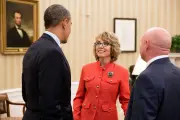 President Barack Obama, wearing a black blazer, faces a woman with a light skin tone wearing a red blazer dress with a silver rose pin and glasses. Also, standing next to President Obama is a man with a light skin tone wearing a dark blue blazer. 