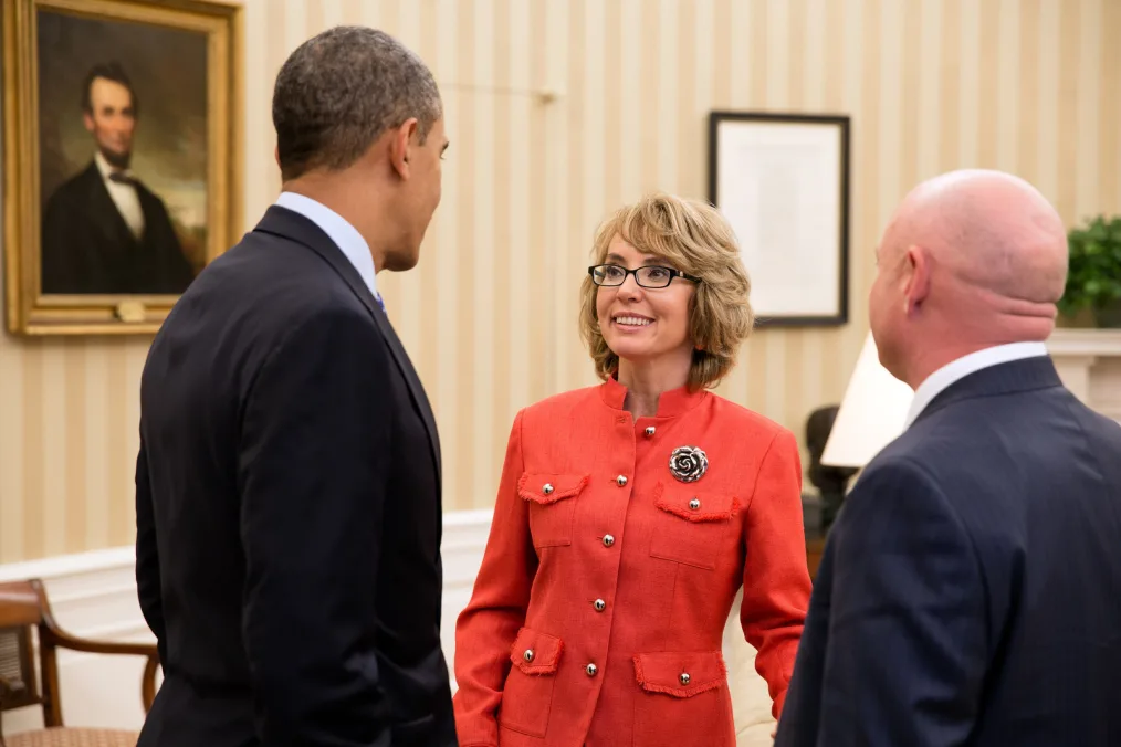 President Barack Obama, wearing a black blazer, faces a woman with a light skin tone wearing a red blazer dress with a silver rose pin and glasses. Also, standing next to President Obama is a man with a light skin tone wearing a dark blue blazer.  