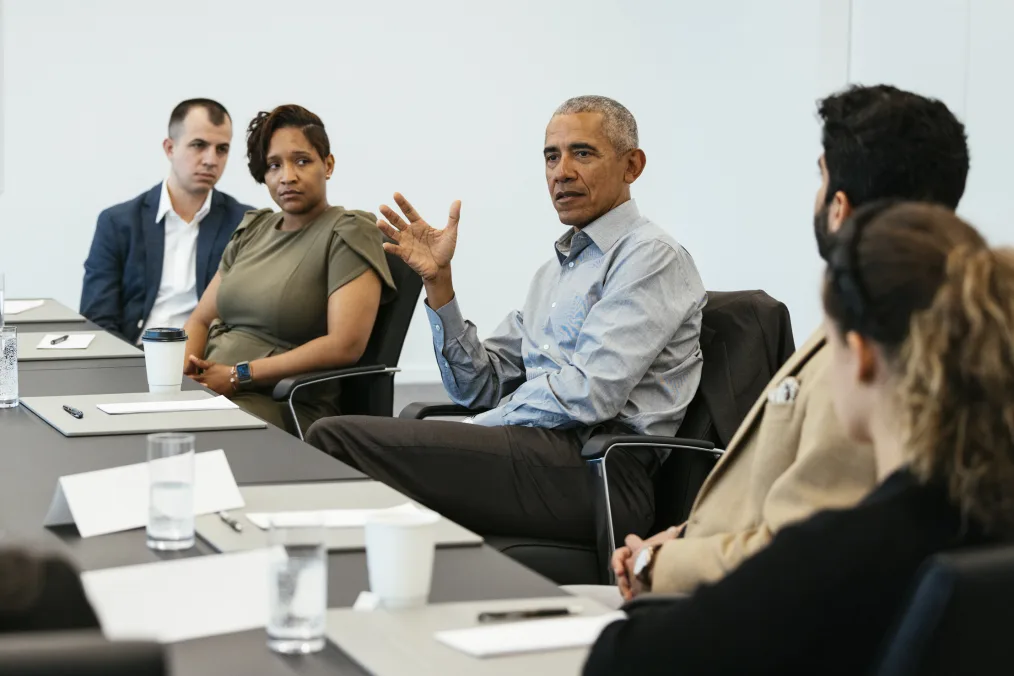 A group of four Obama Scholars dressed in business casual attire listen attentively President Obama at a table during a roundtable meeting. President Obama gestures with his an open palm while speaking.
