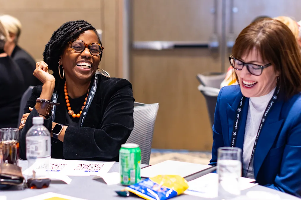 A woman with a dark skin tone sits next to a woman with a light skin tone and brown hair. All are smiling and wearing glasses. 