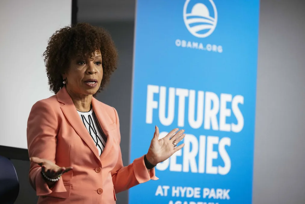 A image of an older woman with medium skin tone. She has a curly brown afro, a peach blazer and a white dress with black lace patterns underneath. She is standing and talking during the Future Series Obama Foundation event.
