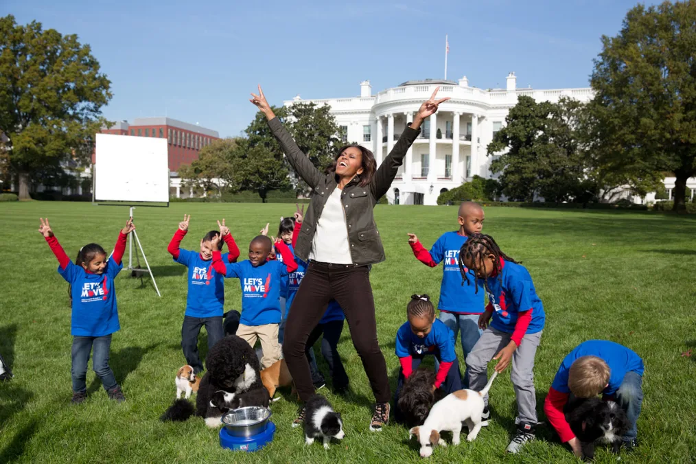 The picture is outside on the lawn of the White House. In the background is the White House. It is sunny outside. Michelle Obama stands in the center of the image, she is smiling with her arms in the air and she is creating a peace sign on both of her hands. She is wearing a dark green jacket, a white shirt and dark pants. Surrounding her are eight children in various stages of movement. The children are all of different races. The children are all wearing blue t-shirts with long sleeved red shirts underneath. On the blue shirts are white text that reads, “Let’s Move!” Around the children are four dogs.