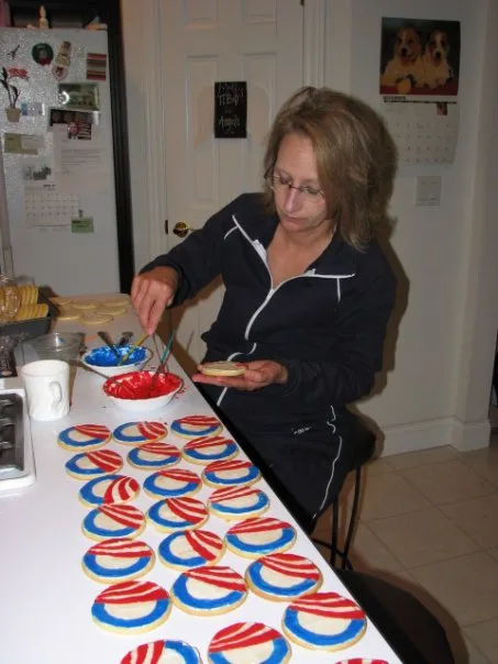 A White woman wearing glasses looks down at her hand as she uses red frosting to decorate blue, red and white cookies in the Obama election campaign "O" logo. There are about 20 cookies on a countertop in front of her.