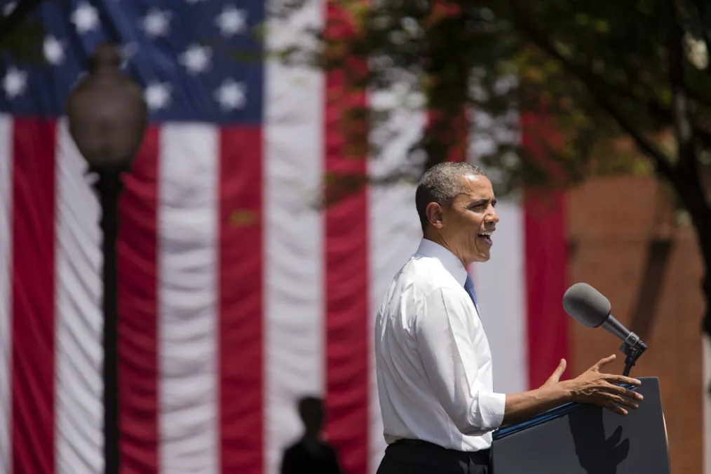 President Obama stands infront of a podium outdoors while giving a speech. A large american flag hangs behind him 