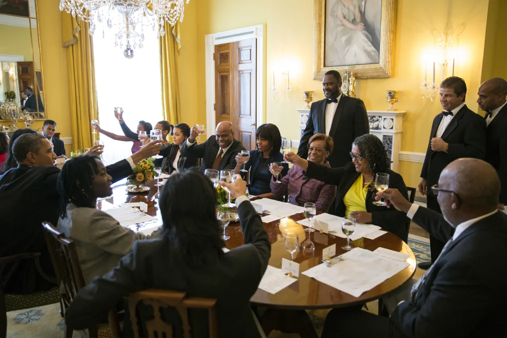 President Obama sits around a table and toasts with a group of people with a range of light and deep skin tones.