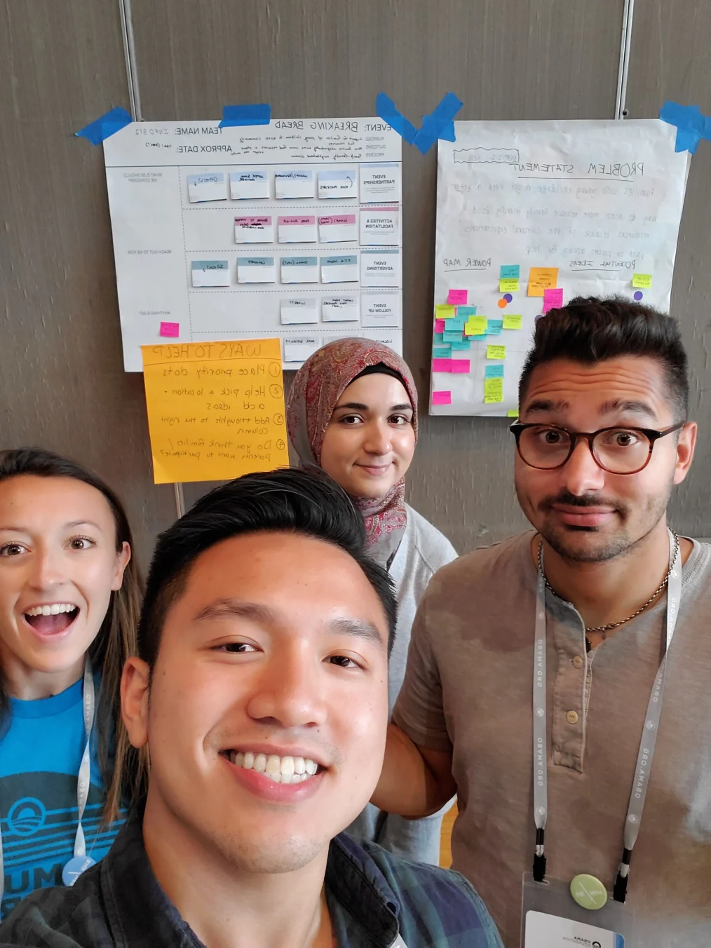 Two women and two men with light-medium skin tones stand in front of two pieces of poster paper with orange, pink, green, and blue sticky notes as they pose for a selfie. 