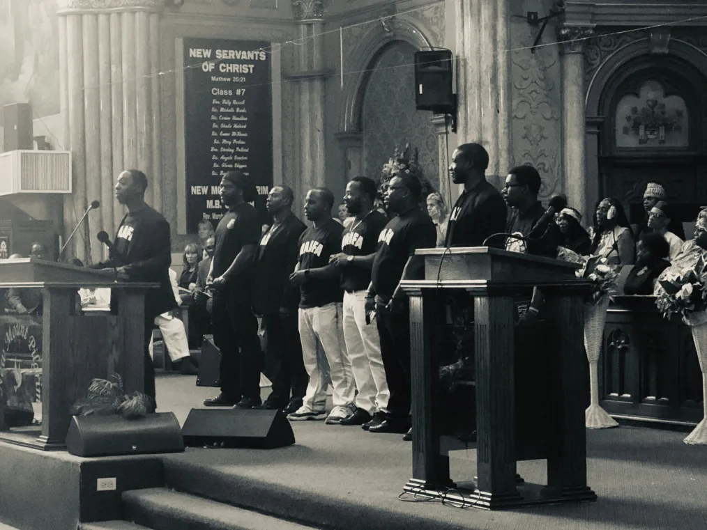 A black-and-white picture focuses on multiple deep skin-toned men standing at church behind the 
speaker at the altar. There is a sign that states "New Servants Of Christ"