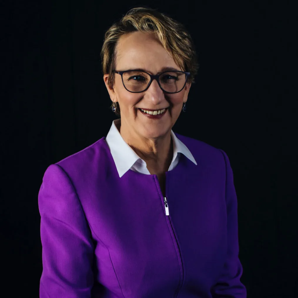 A headshot of a light-skinned woman with short, blonde hair and glasses smiling with her top row of teeth visible. She wears a purple zip-up jacket with a white collar.