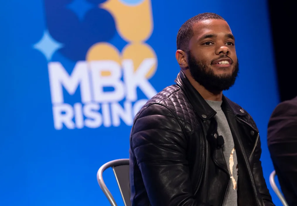 A picture of a young man with medium skin tone, waves, a fade, and a medium sized black beard. He is smiling, looking off slightly to right. The young man is in focus in front of a wall with a design that reads MBK Rising.