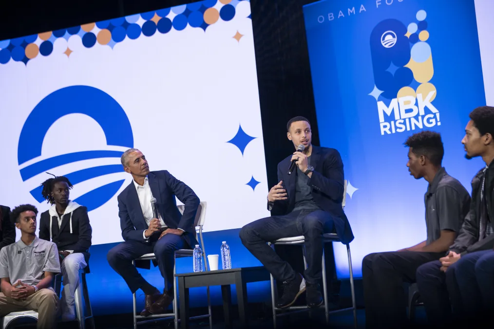 President Obama and Stephen Curry, a man with a light-medium skin tone, light brown hair, wearing a dark blue suit jacket and jeans, speak to a panel of young men with various skin tones. The background includes white, orange, and blue ¨MBK Rising¨ and Obama Foundation graphics.