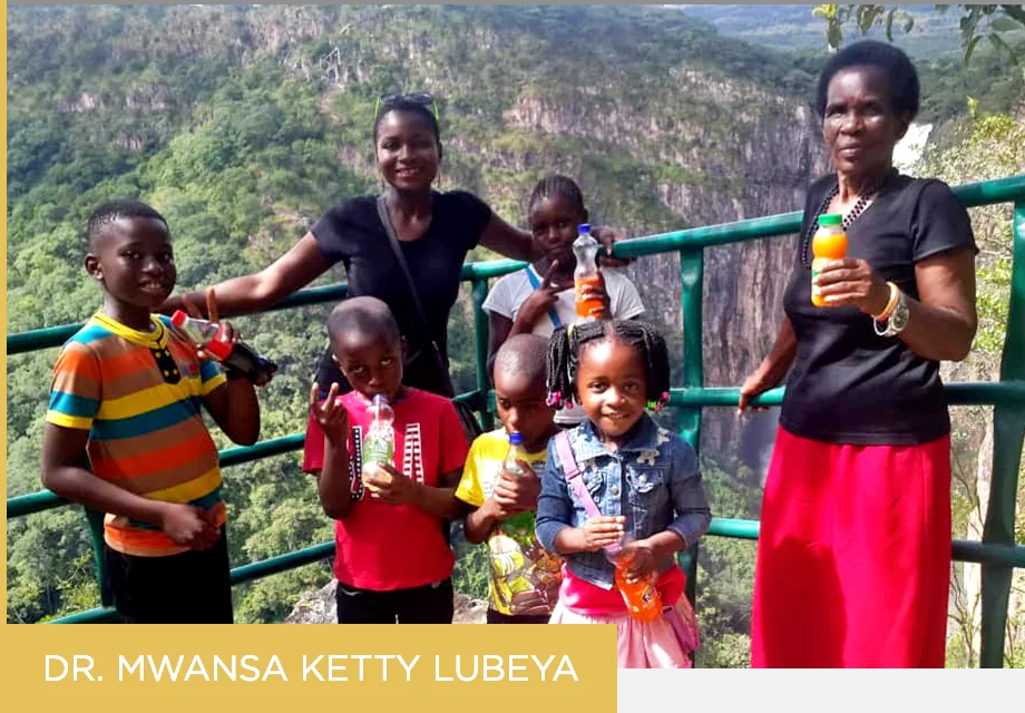 A group photo of young kids and women with deep skin tones stands together as they take a picture in front of large green mountain rocks. Below the picture, a text reads "Dr. Mwansa 
Ketty Lubeya" 