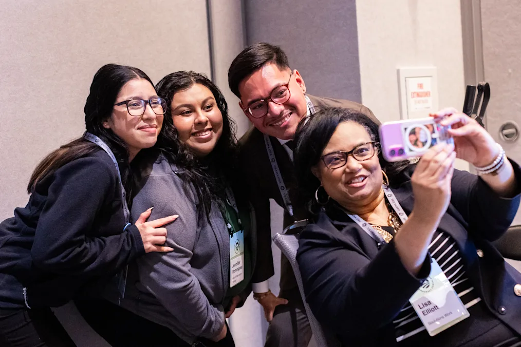 A group of people with a range of light and medium skin tones gather for a selfie. They are all dressed professionally.