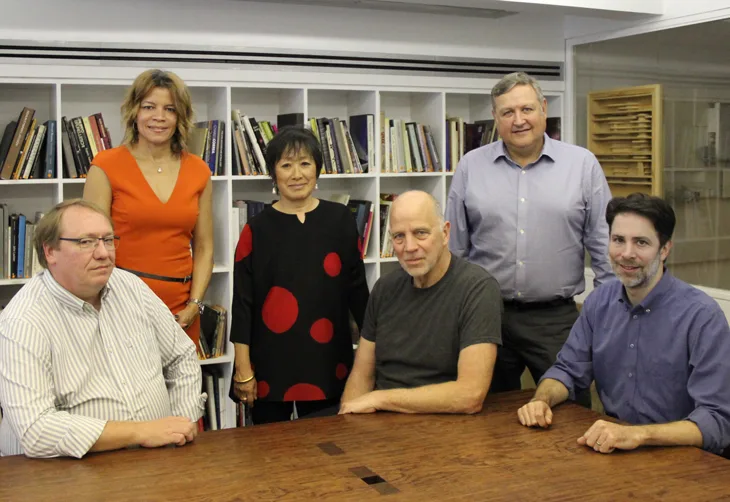Four men and two women surround a wood table and pose for the camera. A wall of books is behind them.