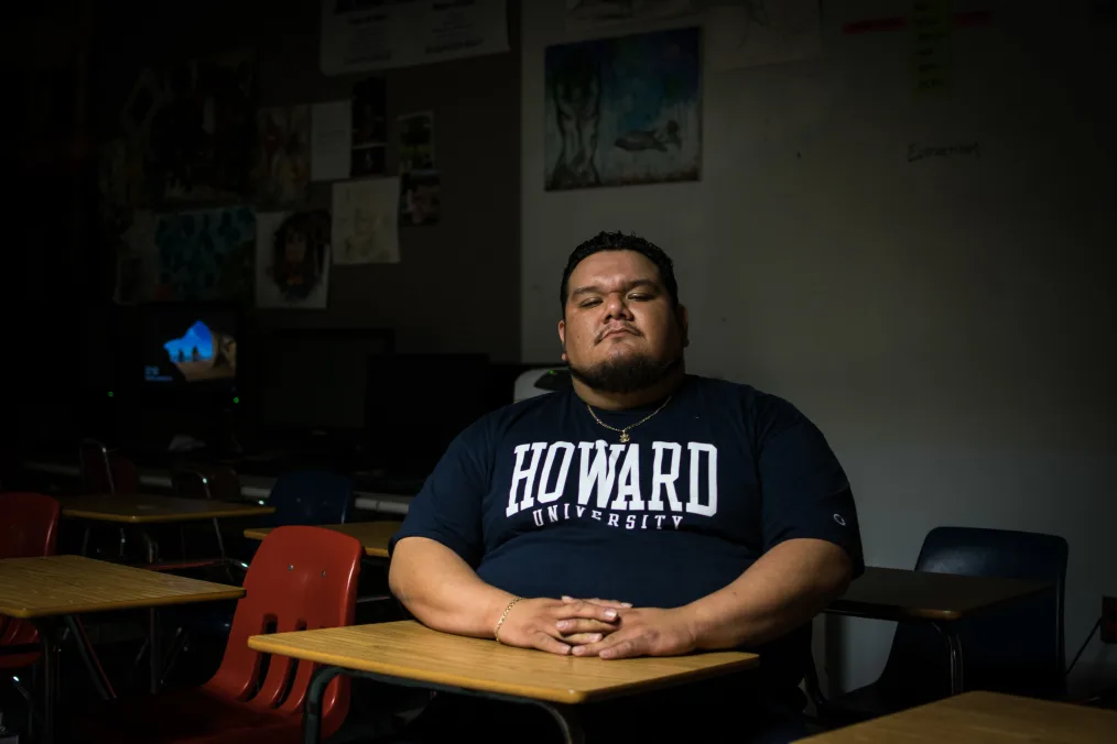 A medium skin toned Brown man with short black hair and a beard is sitting at a desk in a dark classroom. He is wearing a T-shirt that reads HOWARD UNIVERSITY, a gold necklace and a silver bracelet on his right wrist. He is facing the camera with a straight expression. 