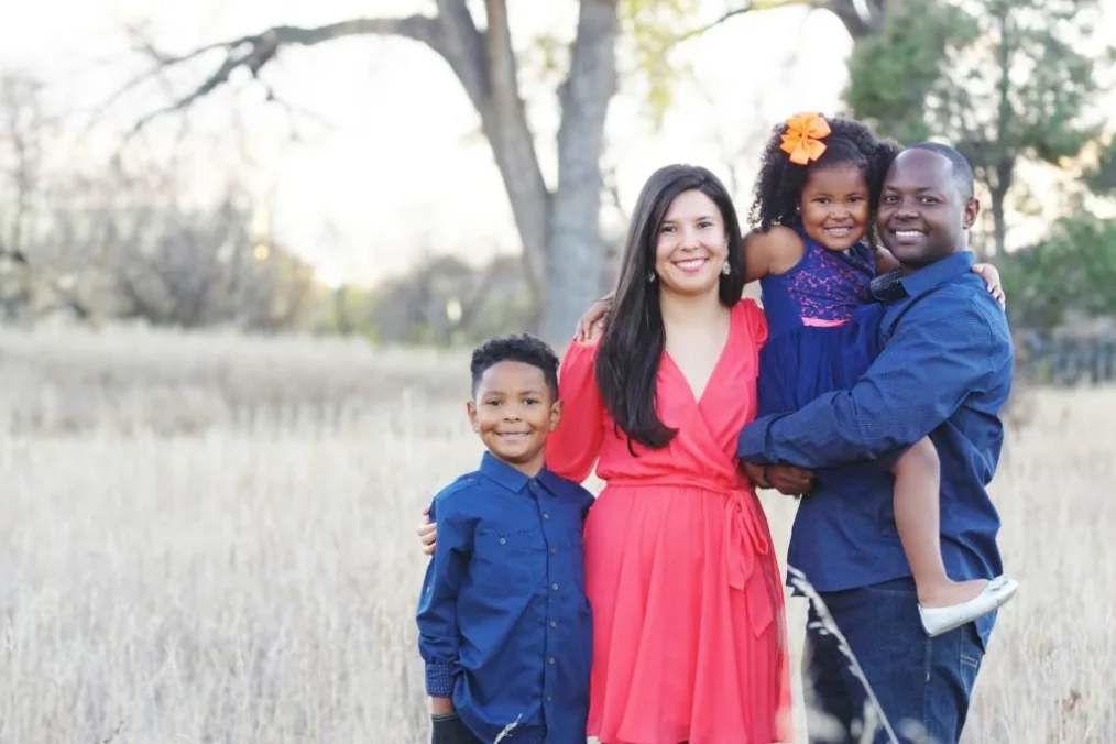 A family of 4 takes a photo standing in open grass. A young boy with a deep medium skin tone and a blue button up, a woman with a medium skin tone and a red drees, a young girl with a deep medium skin tone wearing a blue dress, and man with a deep skin tone also wearing a blue button up. 