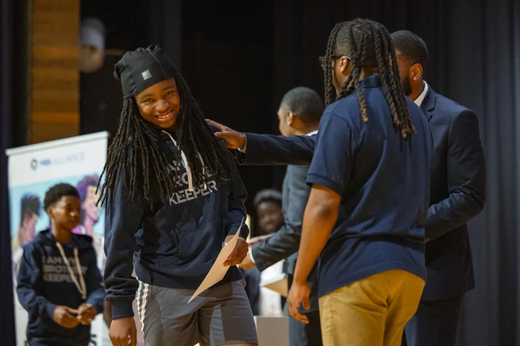 An image of an almond skin toned young man with arm-length locs smiling and leaning slightly to his right as he holds a certificate from another deep skin tone man with other young men and a banner visible in the background. 