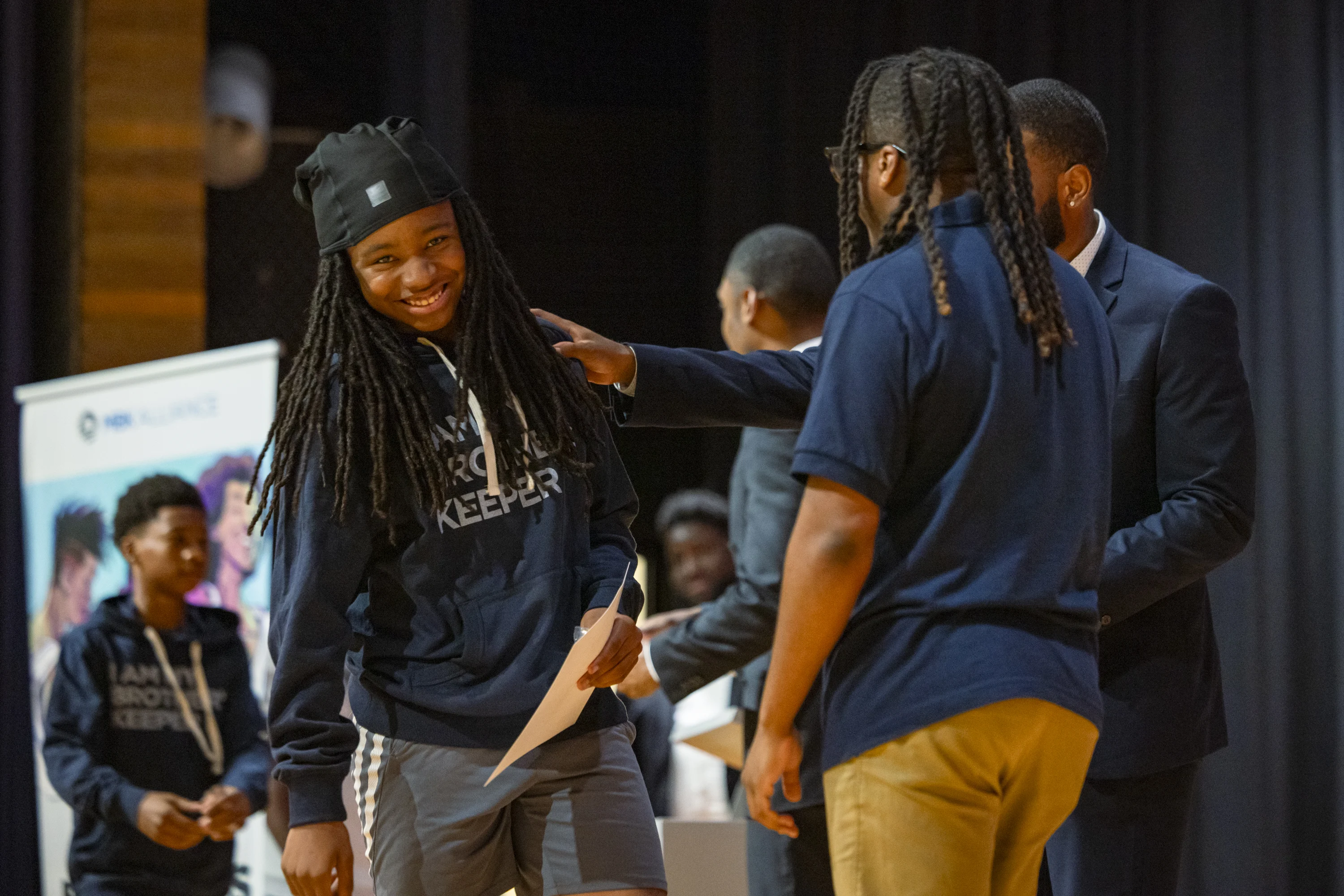 An image of an almond skin toned young man with arm-length locs smiling and leaning slightly to his right as he holds a certificate from another deep skin tone man with other young men and a banner visible in the background. 