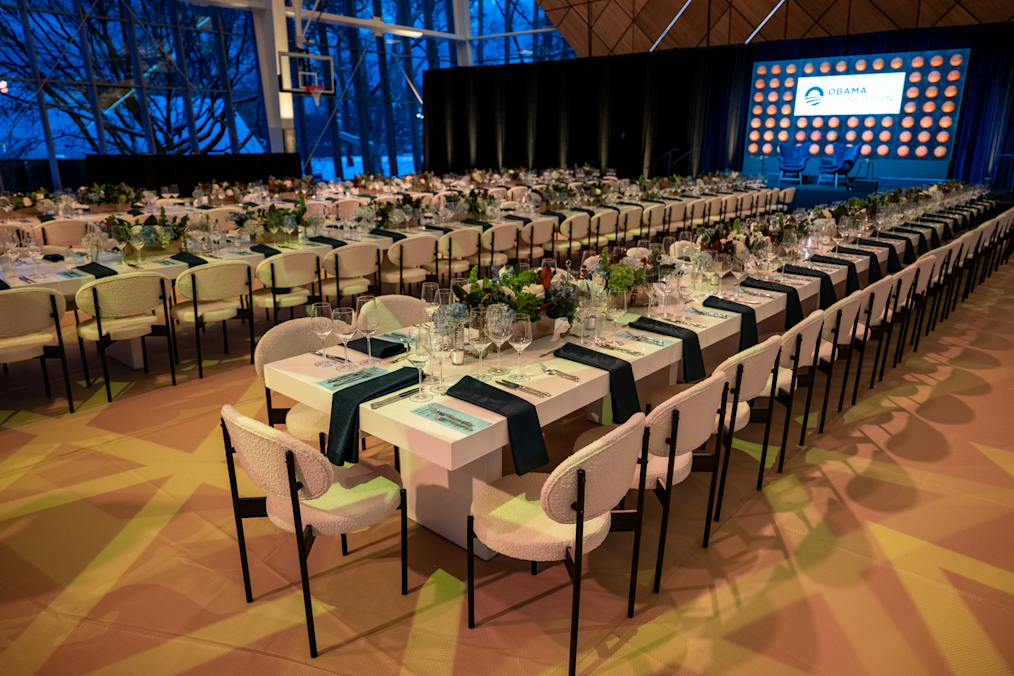 Long white banquet tables lined with cream-colored chairs are set for a formal meal in a large event room. Large windows on the left show a dark blue evening sky outside. At the far end, a stage features a screen with the Obama Foundation logo.
