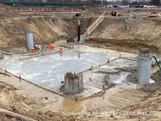 A construction site filled with mud and concrete. Large metal tubing is on either side and a wood staircase is in the back center of the frame.