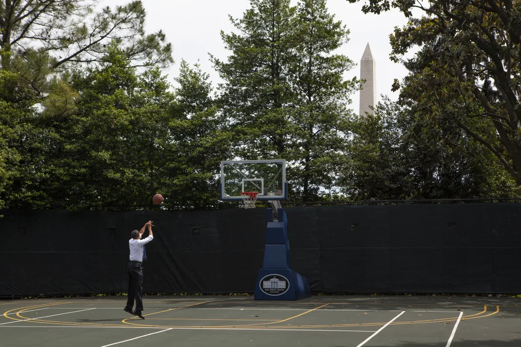 President Obama shoots a basketball into a hoop. He is dressed professionally and is on the White House Basketball court. A White House logo is on the base of the basketball hoop.